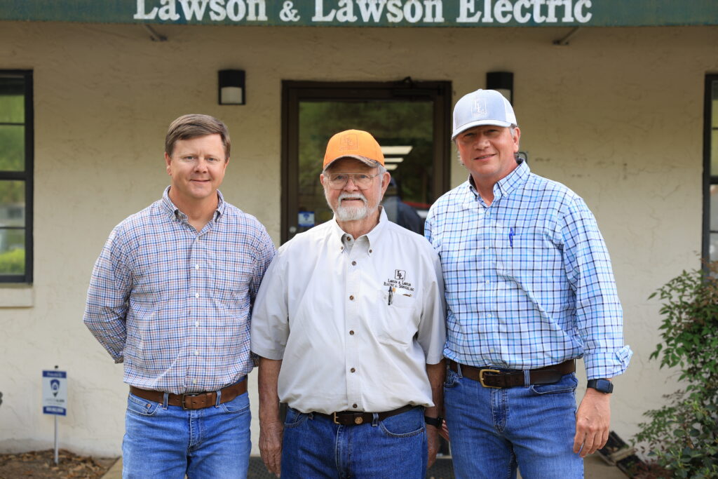 Three men in plaid shirts and caps stand outside an electrical company storefront, smiling at the camera.