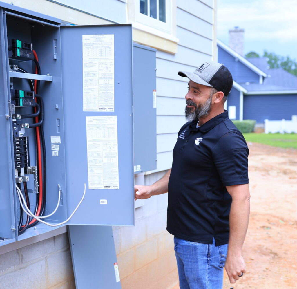 Man wearing a black polo and cap standing beside an open outdoor electrical panel on a house, examining the wiring inside.