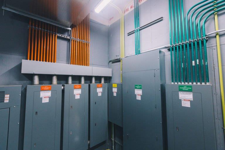 Electrical equipment room with tall gray panels and orange and teal conduit pipes along the wall.