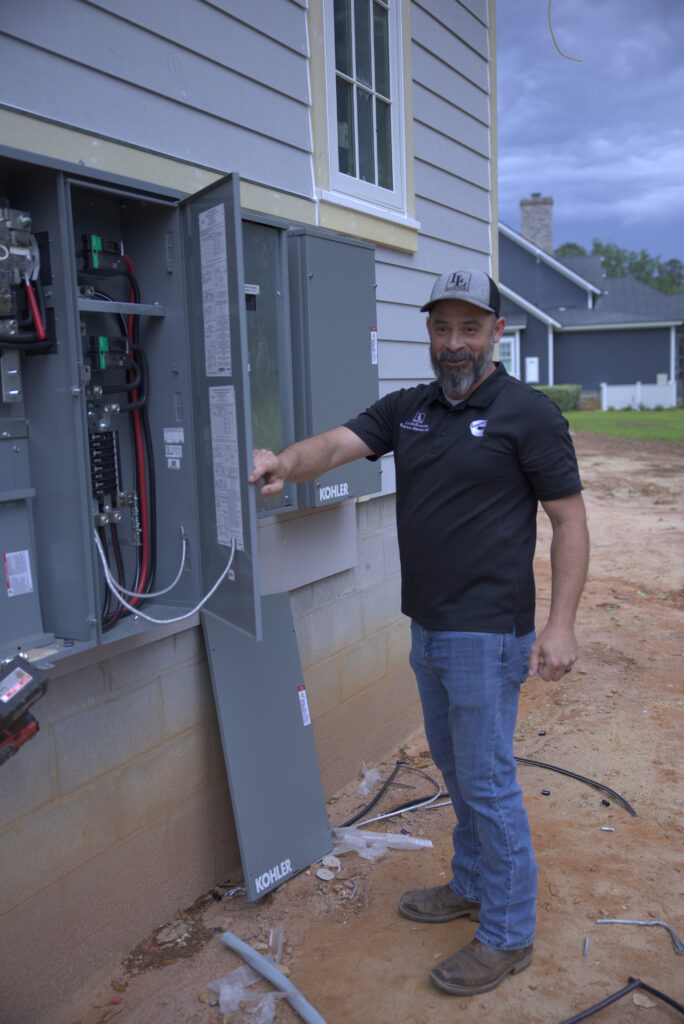 Man in a black polo and cap standing beside an open electrical panel on a house, with construction debris on dirt ground nearby.