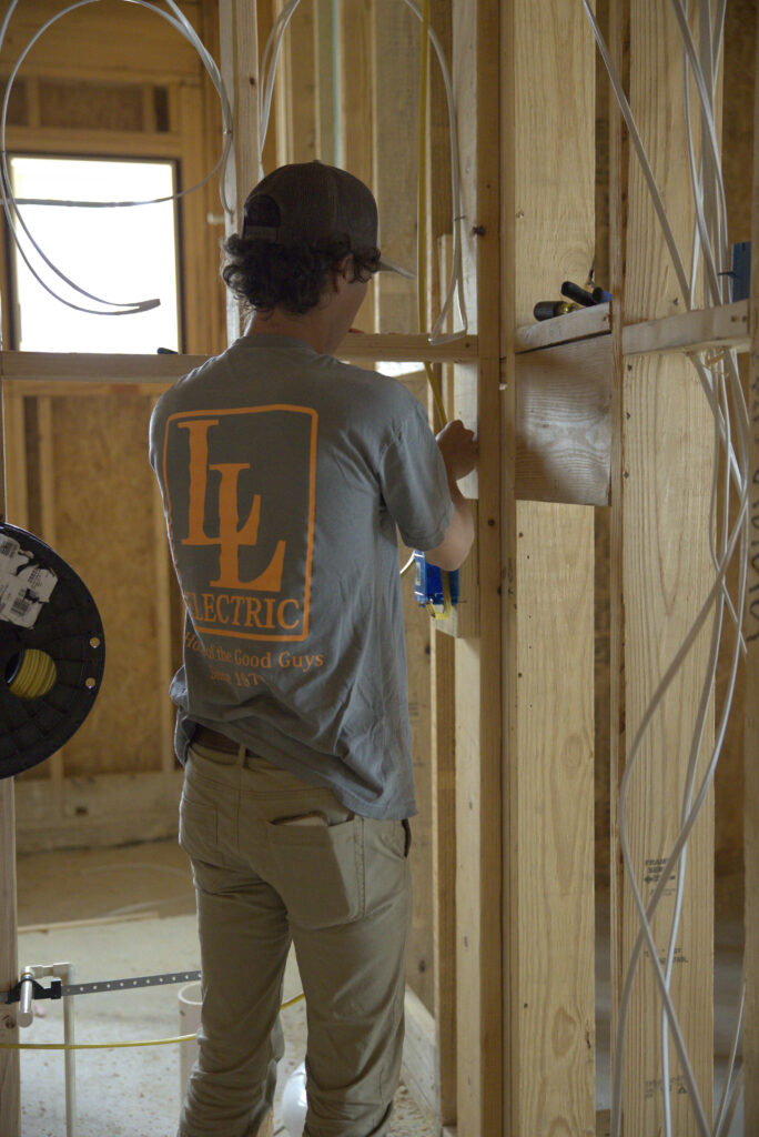 Construction worker in a gray shirt and cap wires a stud wall in a partly finished room, with cables around him.